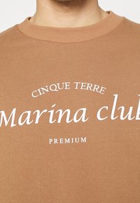 Zbliżenie na brązową bluzę z okrągłym dekoltem i białym napisem „Cinque Terre Marina club Premium” na piersi.