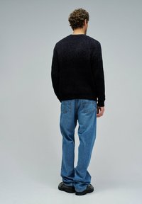 Uomo in piedi di spalle che indossa un maglione nero con texture, jeans blu larghi e scarpe nere, contro uno sfondo grigio chiaro uniforme.