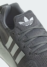 Close-up of gray Adidas sneaker with knitted texture, white three stripes on side, laces, and Adidas trefoil logo on the tongue.