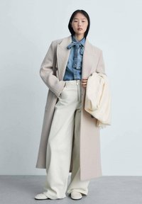 Cappotto in lana beige chiaro sopra una camicia di denim, abbinato a pantaloni bianchi a vita alta e scarpe slip-on color crema. Tiene una sciarpa leggera.