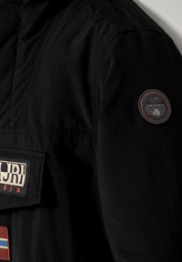 Schwarze Stoffjacke mit großem Logo-Patch und Etikett auf der Brusttasche, mit einem runden Patch-Akzent auf dem Ärmel.