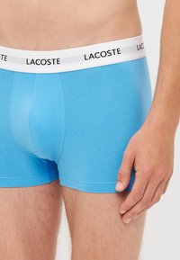 Boxers en coton bleu clair avec une taille blanche portant l'inscription "LACOSTE" imprimée en noir. Texture lisse et design ajusté.