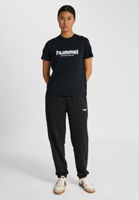 Sort bomulds t-shirt med "hummel" logo i hvid, parret med sorte sweatpants. Fodtøj har beige og sorte accenter. Ensfarvet baggrund.