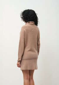 Jasnobrązowy dziergany sweter z wysokim kołnierzem i ściągaczami, w połączeniu z pasującą mini spódnicą z bocznym rozcięciem. Miękka tekstura.