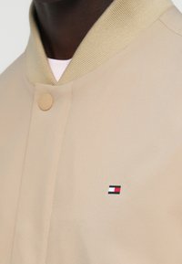 Μπεζ bomber μπουφάν με ριμπ γιακά, διαθέτει στρογγυλό κούμπωμα με κουμπιά και ένα μικρό κεντημένο λογότυπο τριών χρωμάτων στο στήθος.