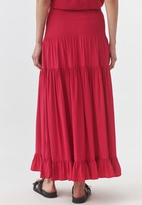 Rode maxi-jurk met smocktaille, voorzien van een gelaagd ontwerp en een gerimpelde zoom. Draagt met zwarte sandalen, weergegeven vanuit de achterkant.