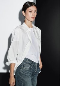 Bílá košile s knoflíky přes bílé tričko, doplněná tmavě šedými džíny. Hladké tkaniny s klasickým límcem, detaily s ohrnutými rukávy a uvolněný střih.
