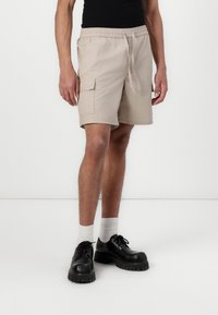 Beige cargo shorts med elastisk talje og snøre, med sidelommer og afslappet pasform. Båret med sorte sko og hvide strømper.