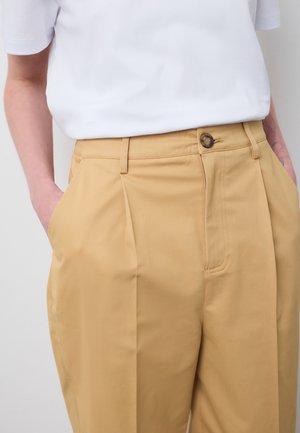 Weißes Kurzarmhemd in beigen, plissierten Hosen eingefasst, die Hände in den Taschen, mit sichtbaren Knöpfen und Gürtelschlaufen an der Taille.