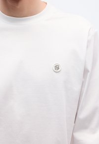 Fehér, hosszú ujjú póló lágy anyagból, kerek nyakkivágással és kontrasztos logófolttal a mellrészen.