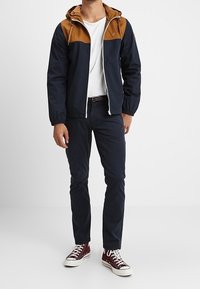 Giacca bicolore blu navy e marrone con cappuccio, zip frontale e polsini elastici. Abbinata a pantaloni blu navy e scarpe bordeaux, con dettagli bianchi.