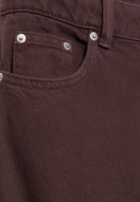 Burgundske traperice z značilnim sprednjim žepom s srebrnim okovjem, teksturirano tkanino in subtilnim diagonalnim tkanjem.