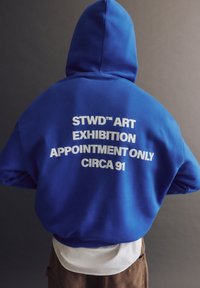 Felpa blu con testo bianco sul retro che recita "STWD™ ART EXHIBITION SOLO SU APPUNTAMENTO CIRCA 91," caratterizzata da una vestibilità comoda e cappuccio.