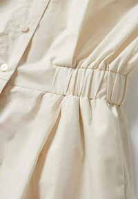 Tela color crema con una textura suave, que presenta botones, fruncido elástico en la cintura y un delicado detalle de drapeado.