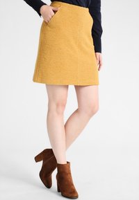 Jupe A-line jaune, texturée, avec poches latérales. Associée à des bottes à chevilles en daim marron. Finition lisse, design mi-long.