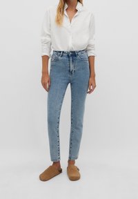 High-waisted light blue denim jeans with a slim fit, five pockets, and a faded texture. Paired with a white button-up shirt and brown clogs.
