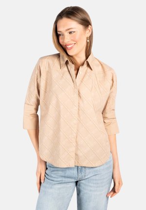 Kvinne smilende, iført beige mønstret skjorte med knapper og trekvart lange ermer, samt lyseblå jeans, stående mot en ensfarget bakgrunn.