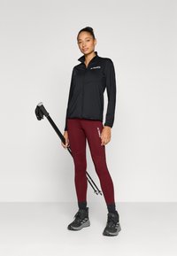 Fekete cipzáros, könnyű dzseki mellzsebbel, bordó leggingsszel párosítva. A modell jobb kezében fekete túrabotot tart.