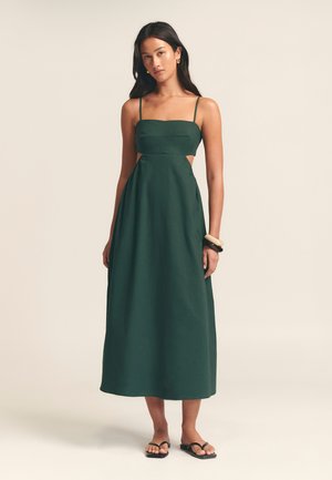 Dark green maxi dress with thin straps, square neckline, cut-out side details, and a smooth, textured fabric. Paired with simple sandals.