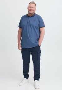 Blaues Polohemd mit dunklem Kragen, marineblaue Cargo-Hosen und weiße Sneakers. Das Material wirkt weich mit gerippten Bündchen und Taschen an den Hosen.
