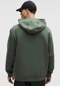 Sudadera con capucha verde de tejido suave, con un corte relajado, puños acanalados y un dobladillo redondeado. La vista trasera muestra la capucha.