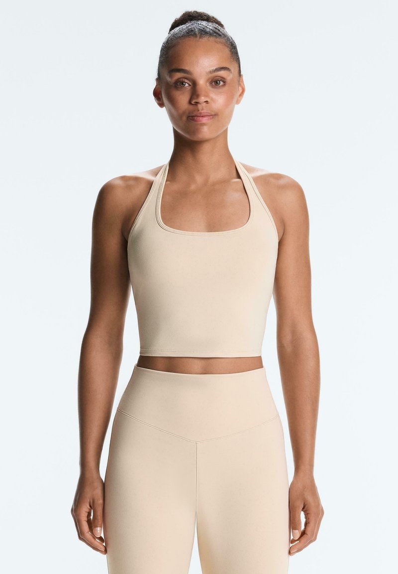 Femme avec les cheveux en chignon, portant un crop top beige à col halter et des leggings taille haute assortis, debout devant un fond clair.