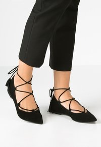 Bailarinas de ante negro con puntera puntiaguda, que presentan cordones entrelazados y detalles de borlas en el tobillo. Diseño de tacón bajo para mayor comodidad.