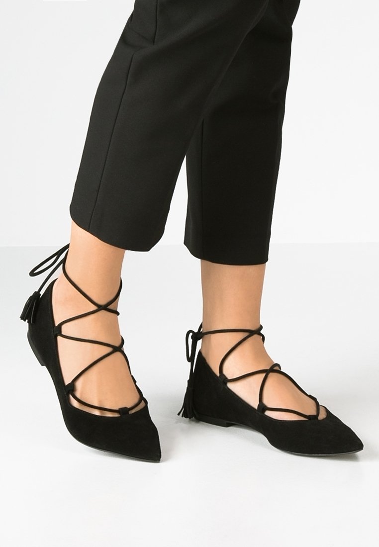 Bailarinas de ante negro con puntera puntiaguda, que presentan cordones entrelazados y detalles de borlas en el tobillo. Diseño de tacón bajo para mayor comodidad.