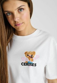 T-shirt en coton blanc avec un graphique d'un ours portant des lunettes de soleil et tenant un téléphone, avec le texte "CERISES" et des contours de cœurs roses.