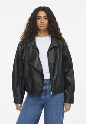 Un mannequin aux longs cheveux bouclés porte une veste en cuir noire sur une chemise blanche et un jean bleu, sur un fond uni.