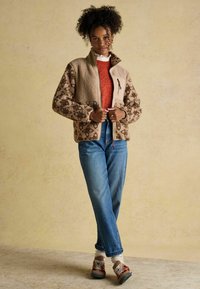 Woman standing with hands adjusting beige floral patterned fleece jacket, wearing orange sweater, blue jeans, and brown sneakers with white socks.