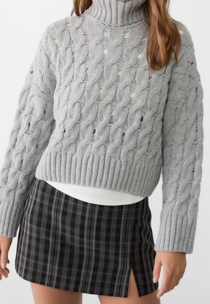 Femme portant un pull à col roulé en maille torsadée gris clair et une mini-jupe à carreaux noirs et gris avec une fente latérale.