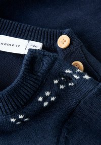 Granatowy sweter dzianinowy z prążkowanym kołnierzem, wyposażony w dwa drewniane guziki oraz białe teksturowane wzory w okolicach dekoltu. Widoczna metka rozmiaru.