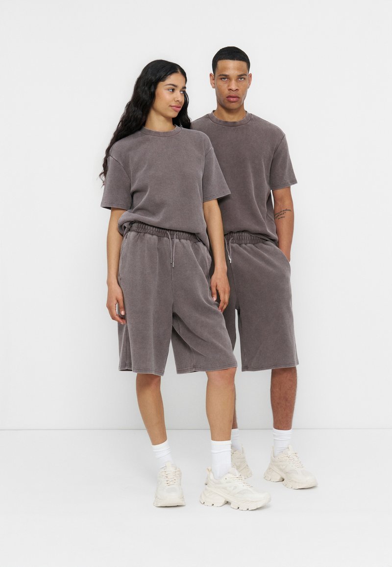 Δύο άτομα φορούν ταιριαστά γκρι oversized T-shirts και σορτς μέχρι το γόνατο με λευκές κάλτσες και chunky sneakers, στέκονται δίπλα-δίπλα.