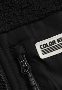 Detailný záber čiernej bundy so zipsom, čiernym a bielym ťahadlom a bielou textilnou etikettou "COLOR KIDS".