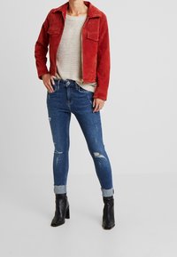 Punainennahkainen takki etutaskuilla, beige neulottu villapusero, kulutettu sininen farkut käännetyillä lahkeilla ja mustat korkeat nilkkakengät.