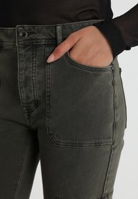 Mörkgröna denimbyxor med en figurnära midja, framficka och synlig sömnad. Tyget har en slät textur och en hållbar design.