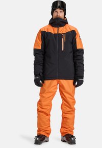 Giacca da sci nera con accenti arancioni, dotata di tasche con cerniera e collo alto, abbinata a pantaloni e guanti arancioni coordinati.