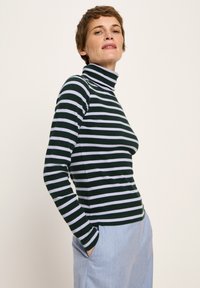 Person in einem grün-weiß gestreiften Rollkragenpullover und hellblauer Hose, der mit den Händen in den Taschen vor einem schlichten Hintergrund steht.