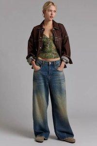Giacca di jeans marrone, top in stile tank verde a fantasia, jeans blu chiari oversize, in piedi con le mani in tasca, calzature marrone chiaro.