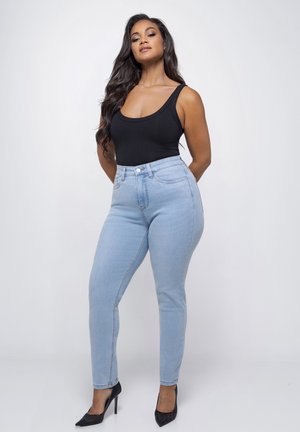 Vrouw met lang donker haar, gekleed in een zwart mouwloos topje, lichtblauwe hoge taille jeans en zwarte hoge hakken, staand tegen een witte achtergrond.