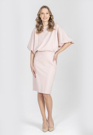 Kobieta o falowanych blond włosach nosi jasnoróżową sukienkę za kolano z szerokimi rękawami oraz beżowe szpilki, stojąc na jednolitym tle.