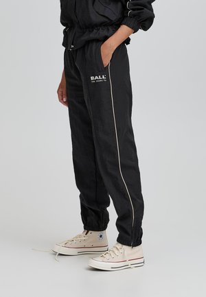 Sorte træningsbukser med en struktureret overflade, der har en hvid side stripe og logoet "BALL" på hoften; parret med cremefarvede høj-top sneakers.