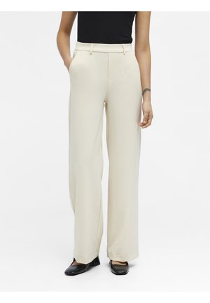 Woman wearing high-waist, wide-leg beige trousers with side pockets and black heeled shoes, standing against a plain white background.