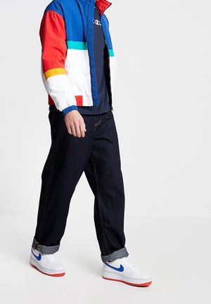 Personne portant une veste coupe-vent color-block, un jean foncé retroussé aux chevilles et des baskets blanches avec un swoosh bleu et des semelles rouges, marchant sur un fond blanc.