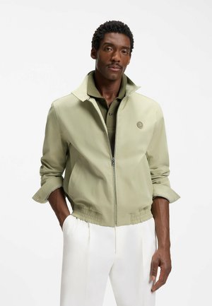 Man met kort krullend haar die een lichtgroene ritstrui draagt over een groen poloshirt en witte broek tegen een effen achtergrond.