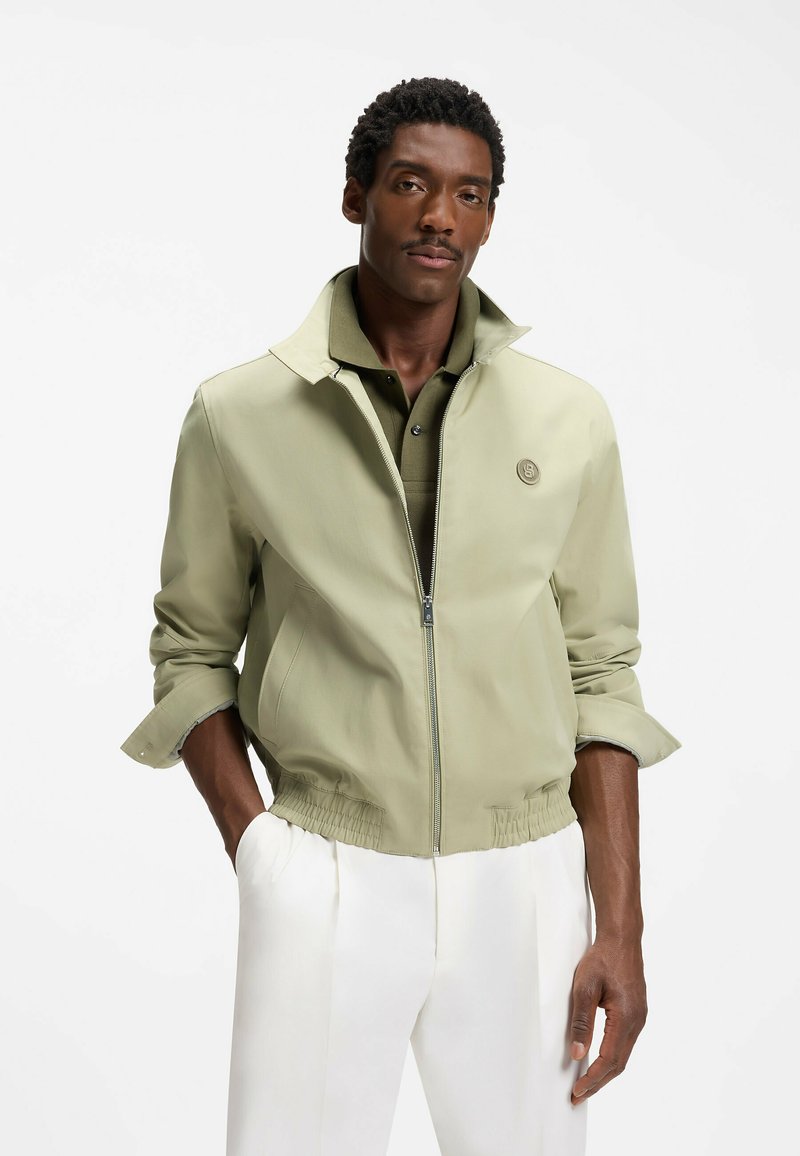 Homme aux cheveux courts et bouclés portant une veste zippée vert clair sur un polo vert et un pantalon blanc, sur un fond uni.