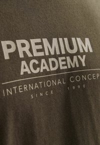 Camiseta de color gris oscuro con texto blanco. El diseño incluye "ACADEMIA PREMIUM" y "CONCEPTOS INTERNACIONALES" con "DESDE 1990" debajo.