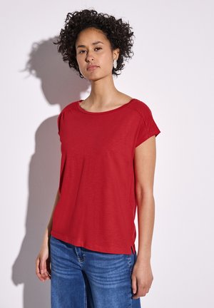 Haut rouge à manches courtes avec un col large, fabriqué en tissu doux, doté de fentes latérales et d'une coupe décontractée. Associé à un jean bleu clair.