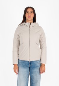 Giacca zip con cappuccio di un beige chiaro, con una texture liscia, tasche laterali e un design aderente, abbinata a jeans blu.